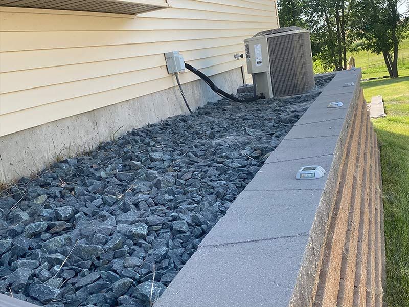 Stone retaining wall with a beige house and air conditioner. Dark rocks cover the ground.