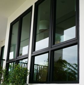 Black-framed windows reflect a building, sky, and plants; white exterior wall; sunny day.