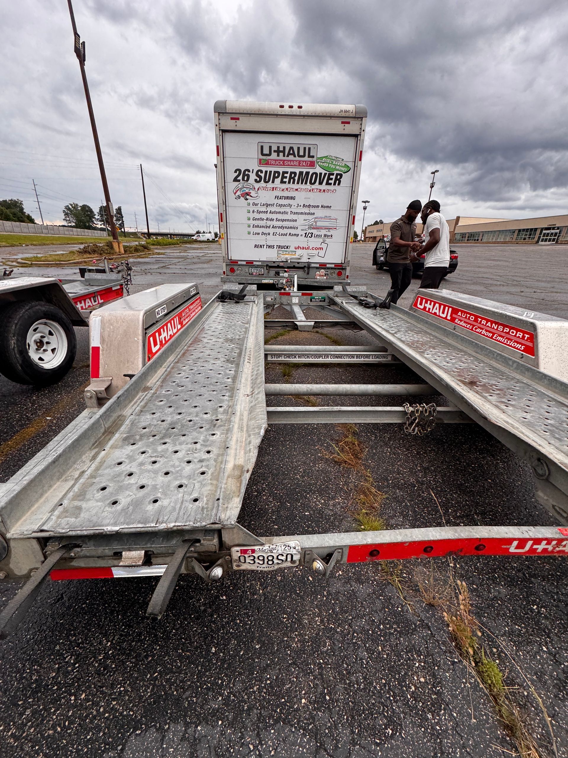 Open car trailer with ramps, two men in the background, cloudy sky.