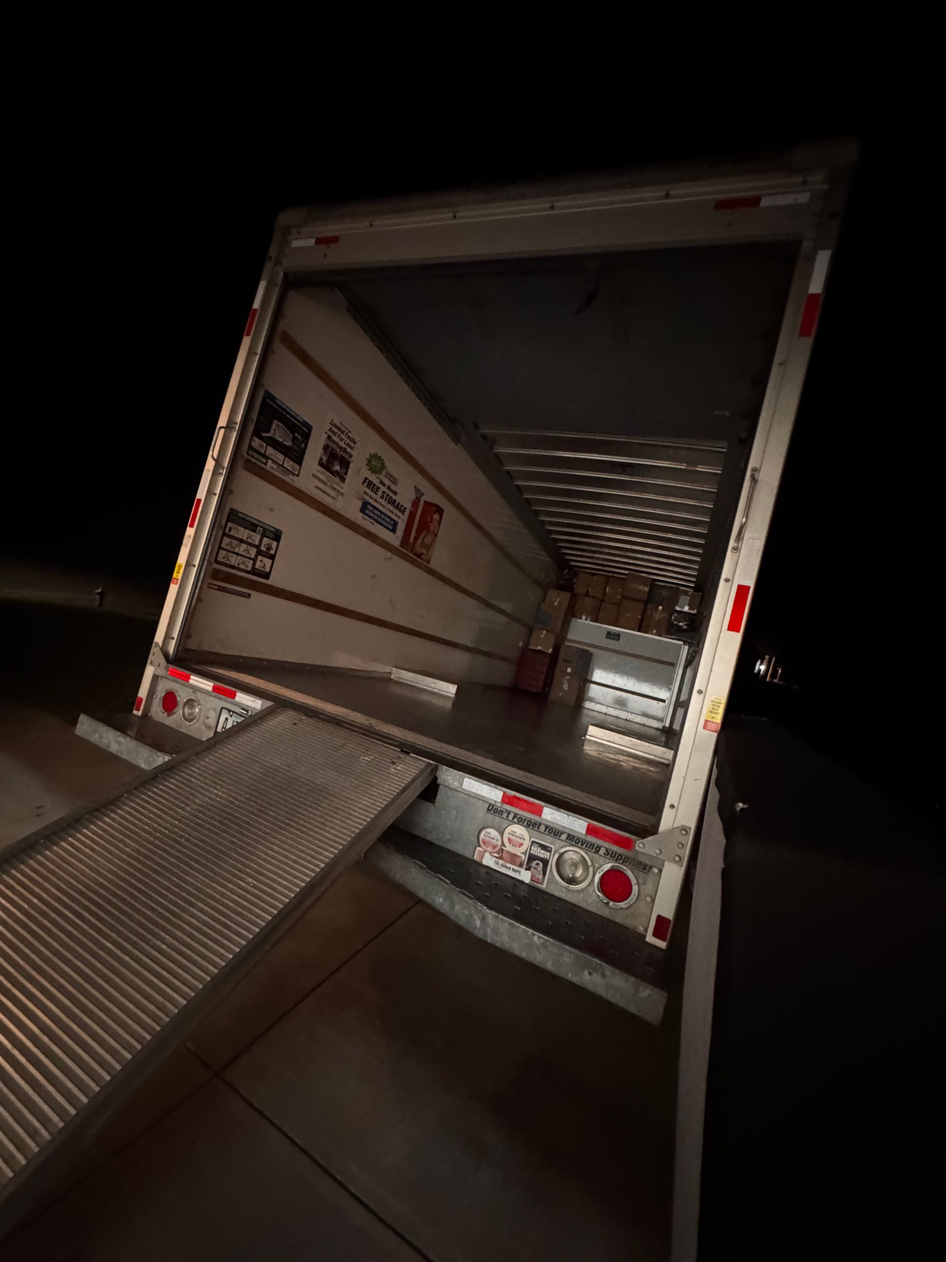Empty semi-trailer cargo area with ramp extended, viewed from ground level in low light.