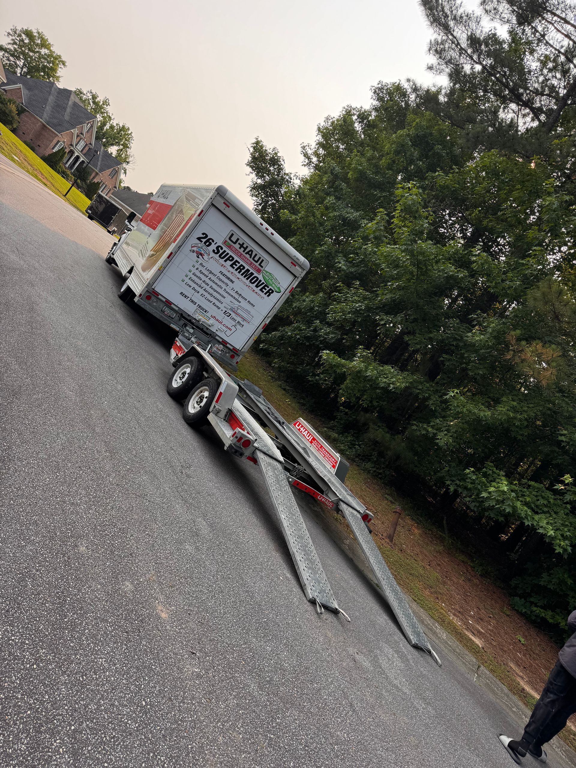 U-Haul trailer on a sloped asphalt road. Two ramps are extended. Trees and a house are in the background.