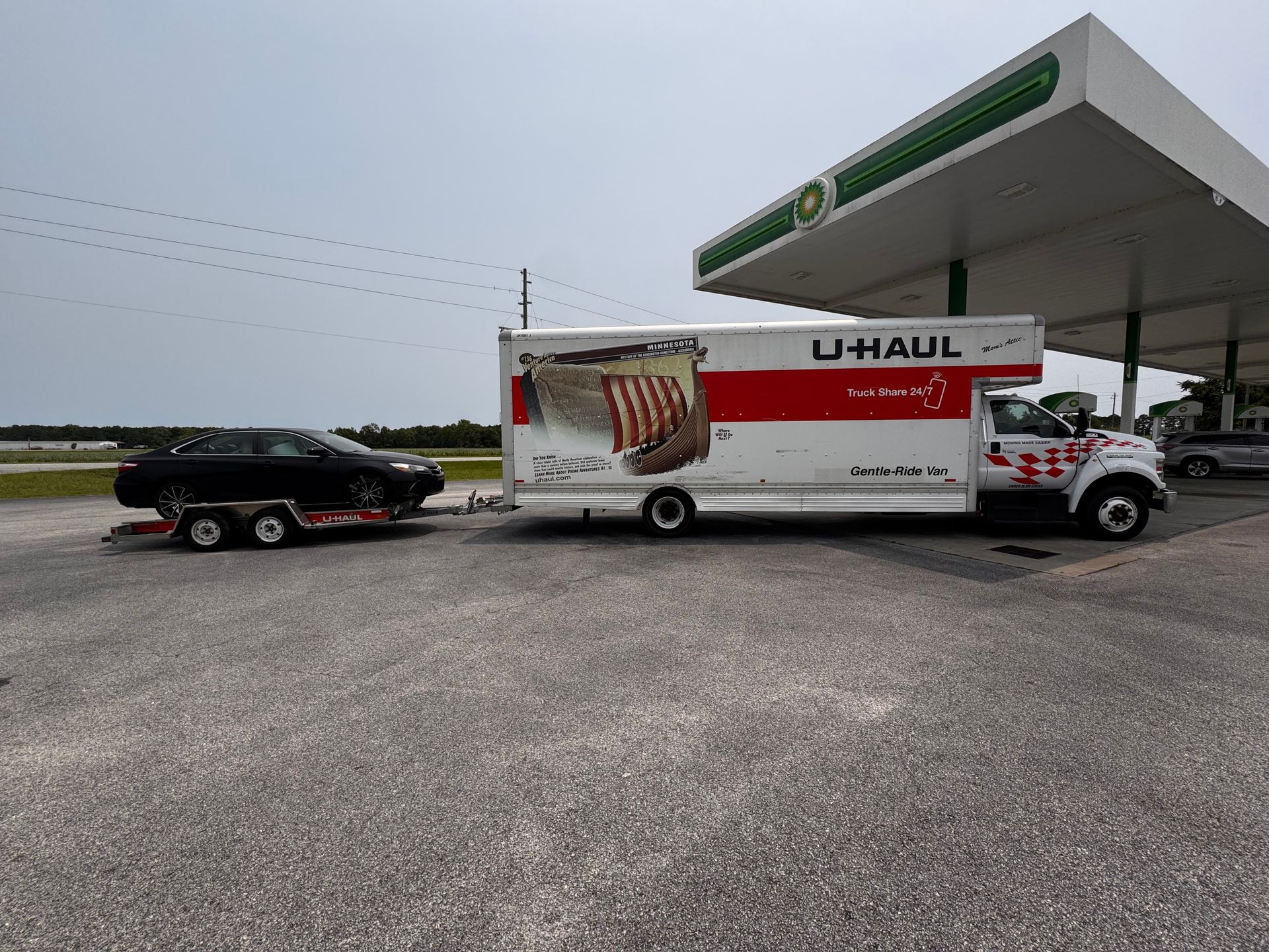 U-Haul truck with a car on a trailer at a BP gas station under a sunny sky.