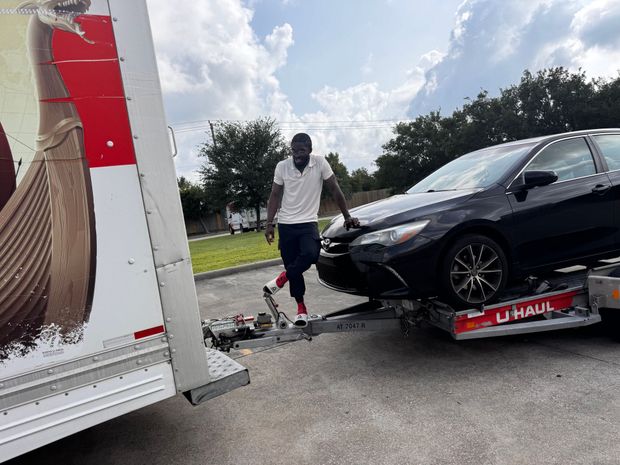 Man standing next to a black car loaded onto a U-Haul trailer.  Bright sky, sunny day.