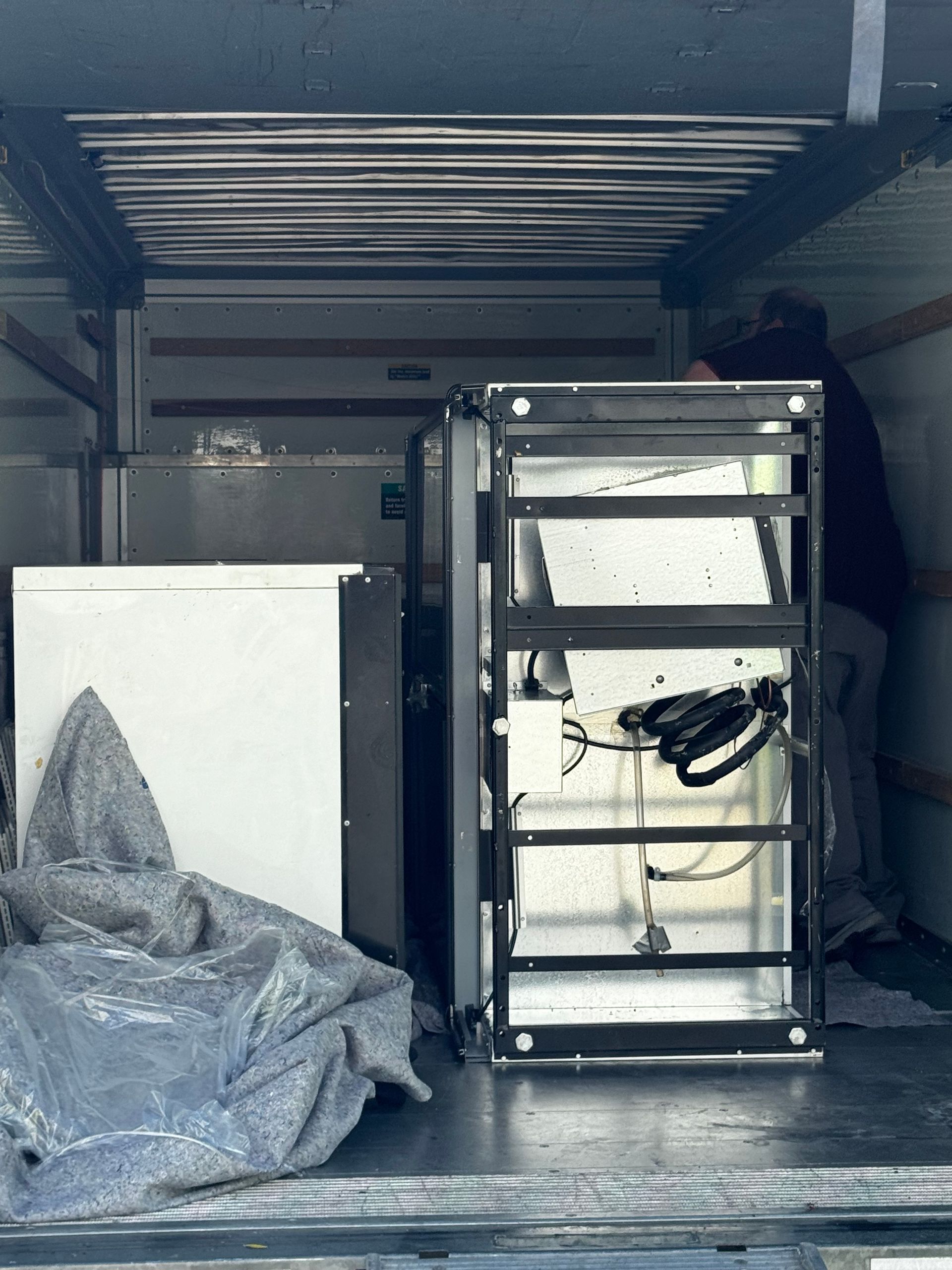 Person in a truck loading appliances: white cabinet, open metal frame, silver machine, and gray tarp.