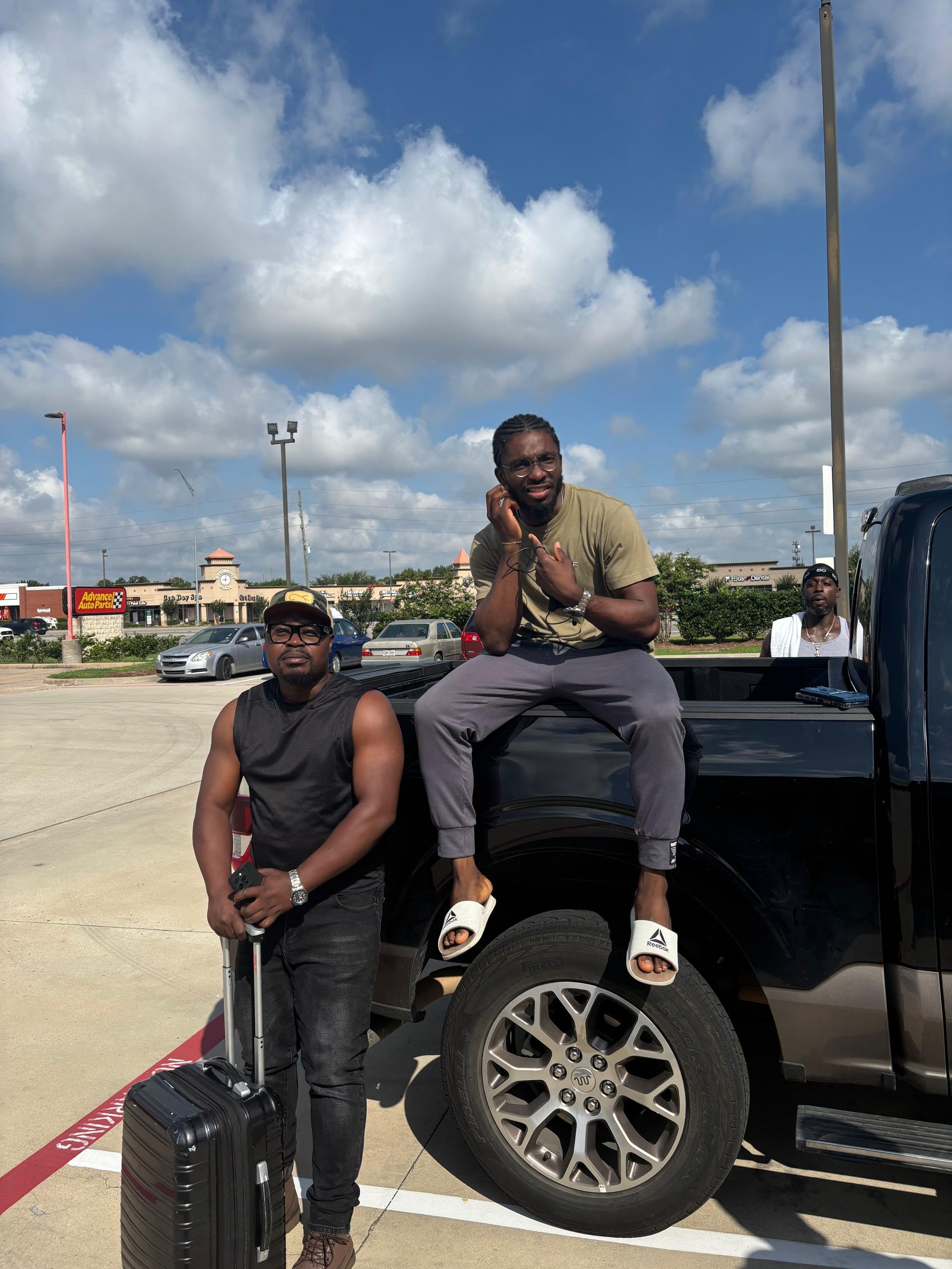 Two men outside; one sits on truck bed, the other holds luggage. Sunny, retail area.