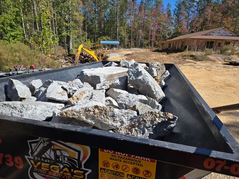 Dumpster filled with concrete rubble at a construction site; excavator and building in the background.