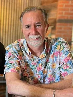 Smiling man in a colorful floral shirt seated indoors with brick wall background