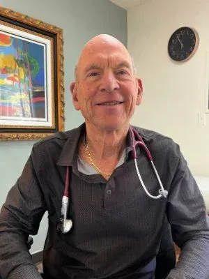 Smiling man in a dark shirt with a stethoscope, seated in an office with framed art and a wall clock.
