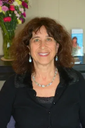Smiling woman in a black top and necklace standing indoors with flowers in the background