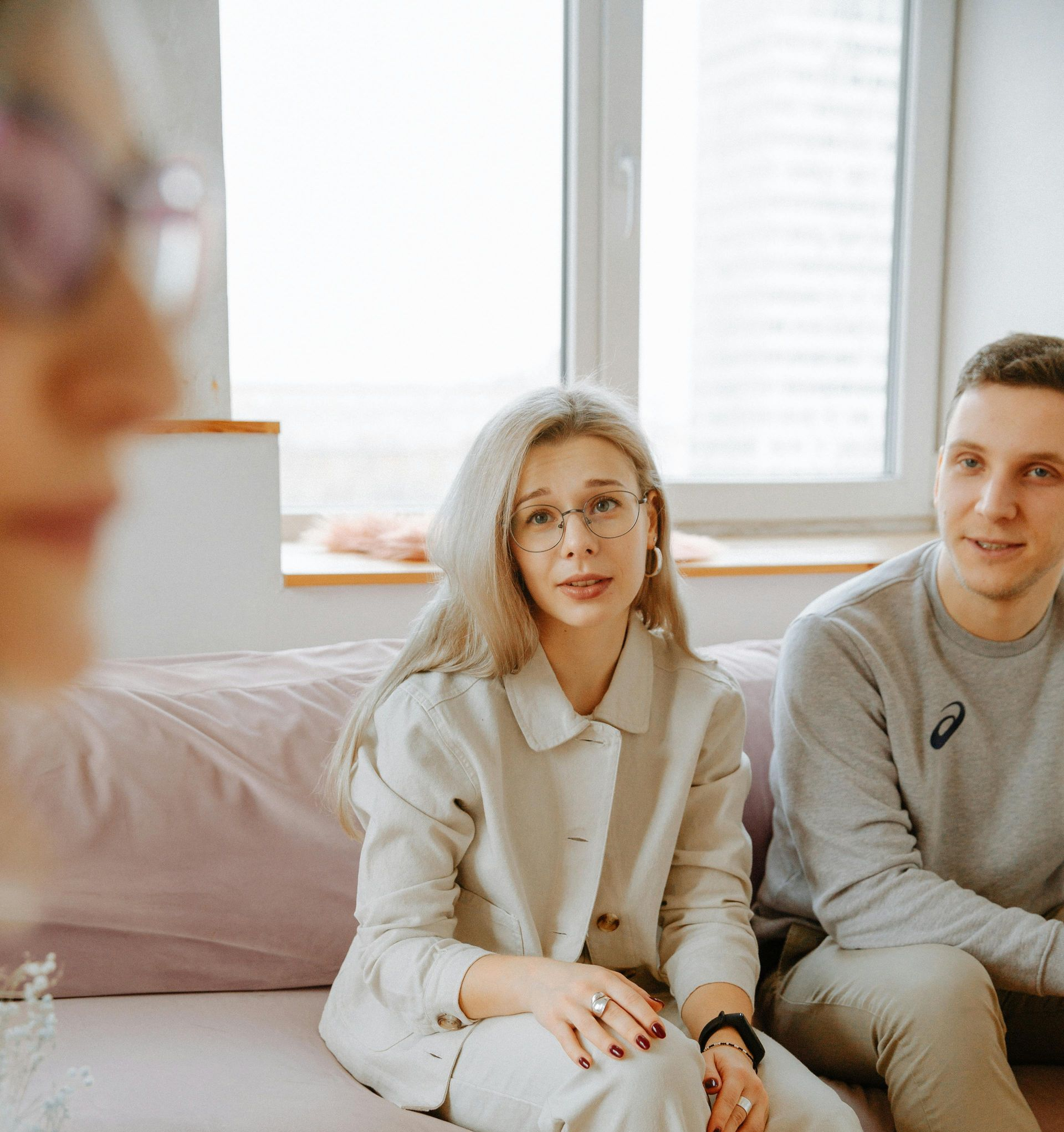 Two people sitting on a sofa in a bright room, looking toward someone off-camera.