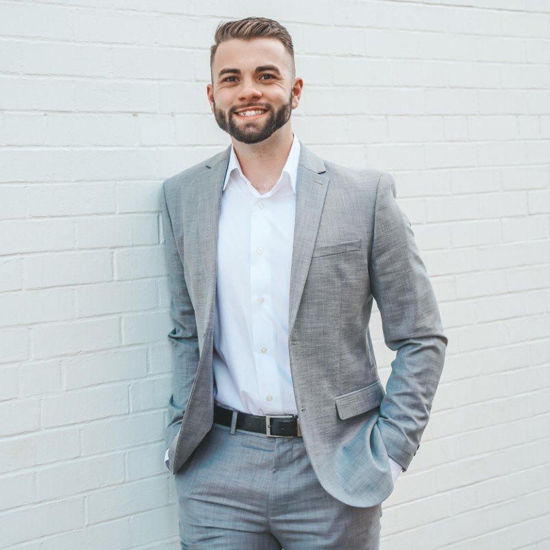 A man in a suit and white shirt is standing in front of a white brick wall.