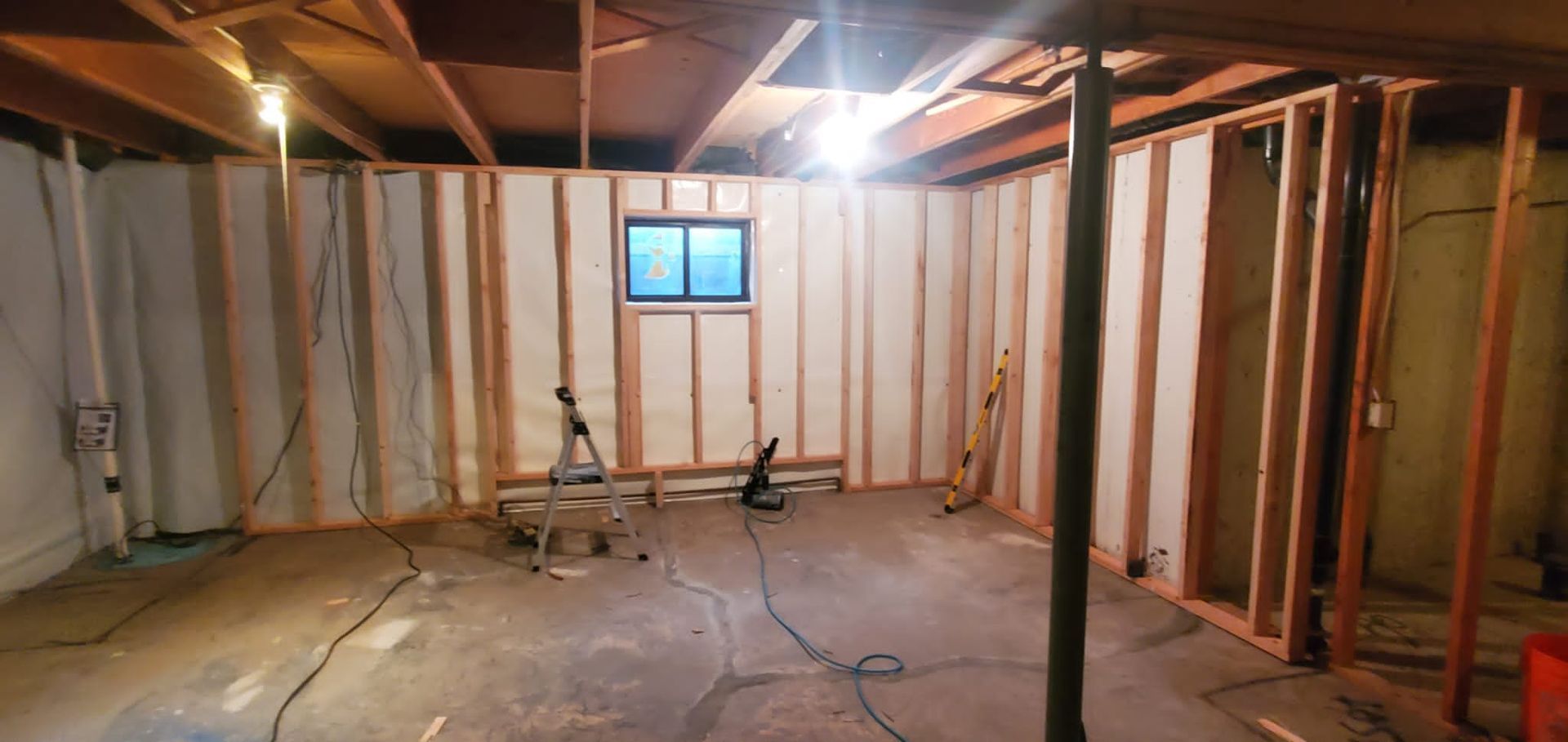 A basement under construction with framed wooden walls, exposed joists, a small window, and a central metal support column.