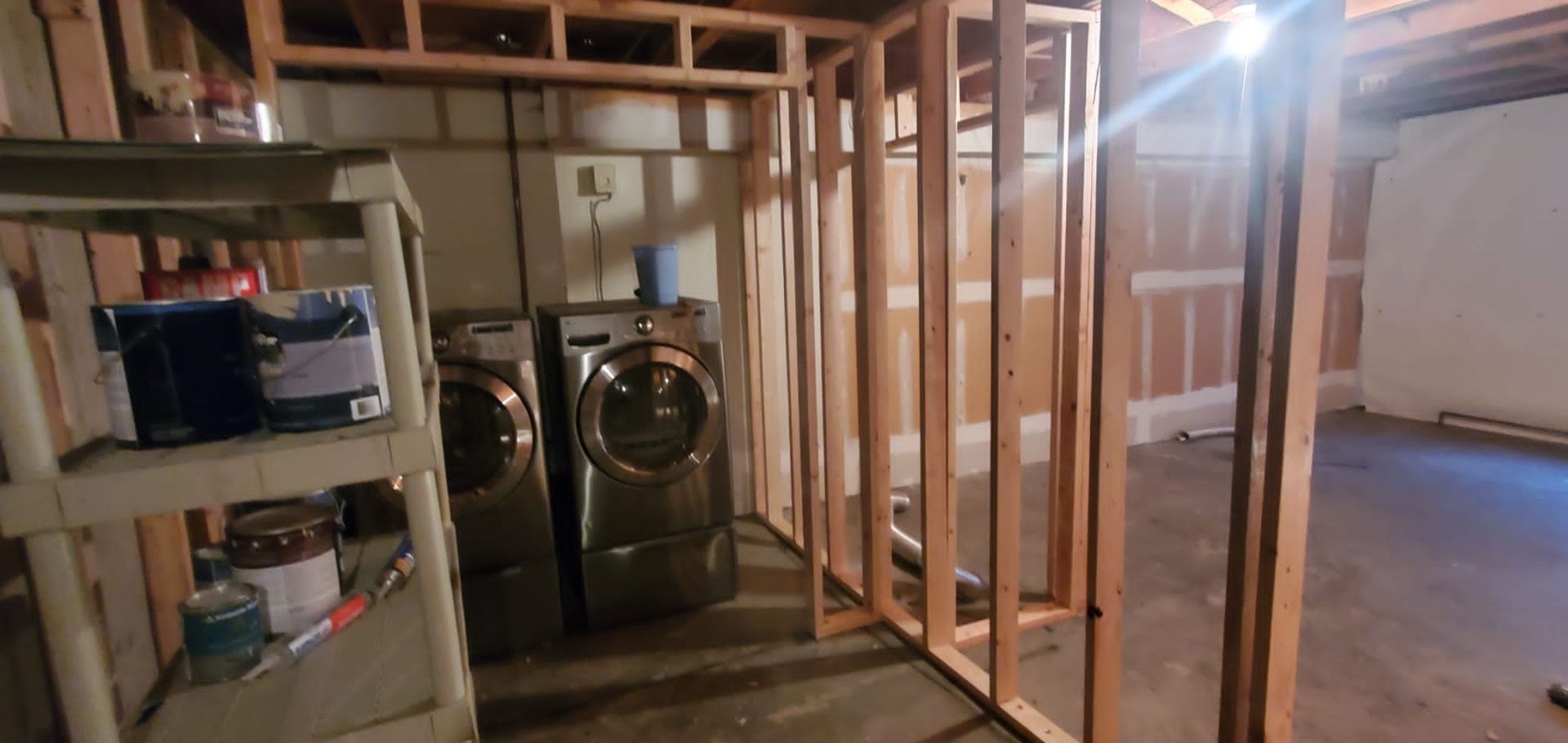 Two laundry machines sit inside an unfinished basement framed with wooden wall studs.