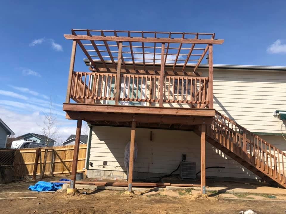 A wooden deck under construction with an overhead pergola frame, attached to the side of a light-colored residential home.