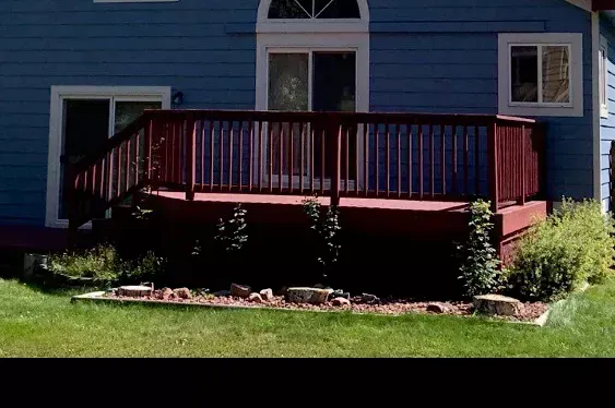 Red wooden deck attached to a blue house with white trim, fronted by a small lawn and shrubs