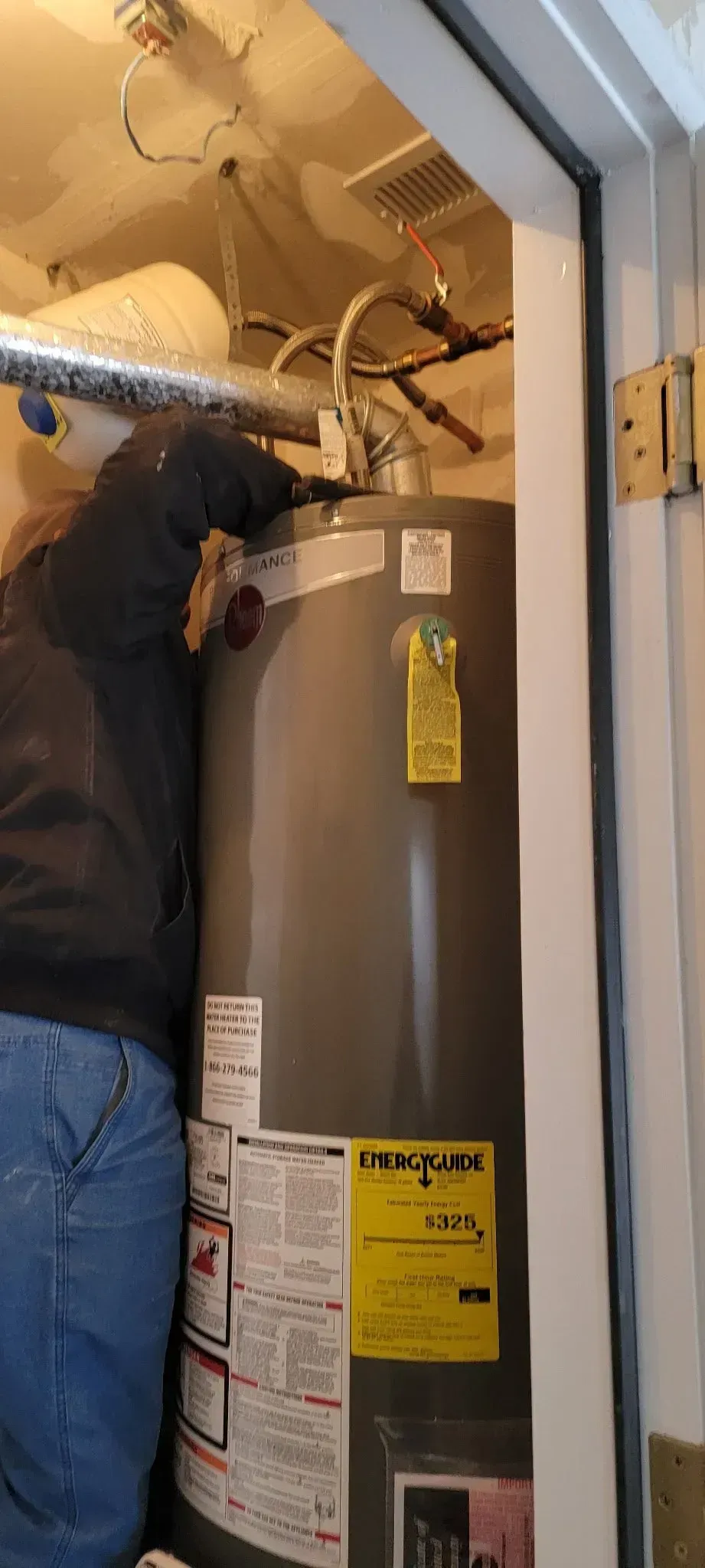 Technician replacing a water heater in a narrow utility closet, with exposed pipes above.