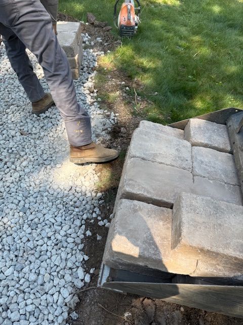 Worker standing on gravel beside stacked paving stones and a grassy area, with an excavator in the background