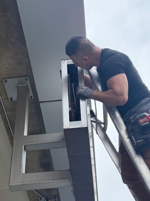 Worker in a black shirt inspecting a wall-mounted HVAC unit from a ladder.