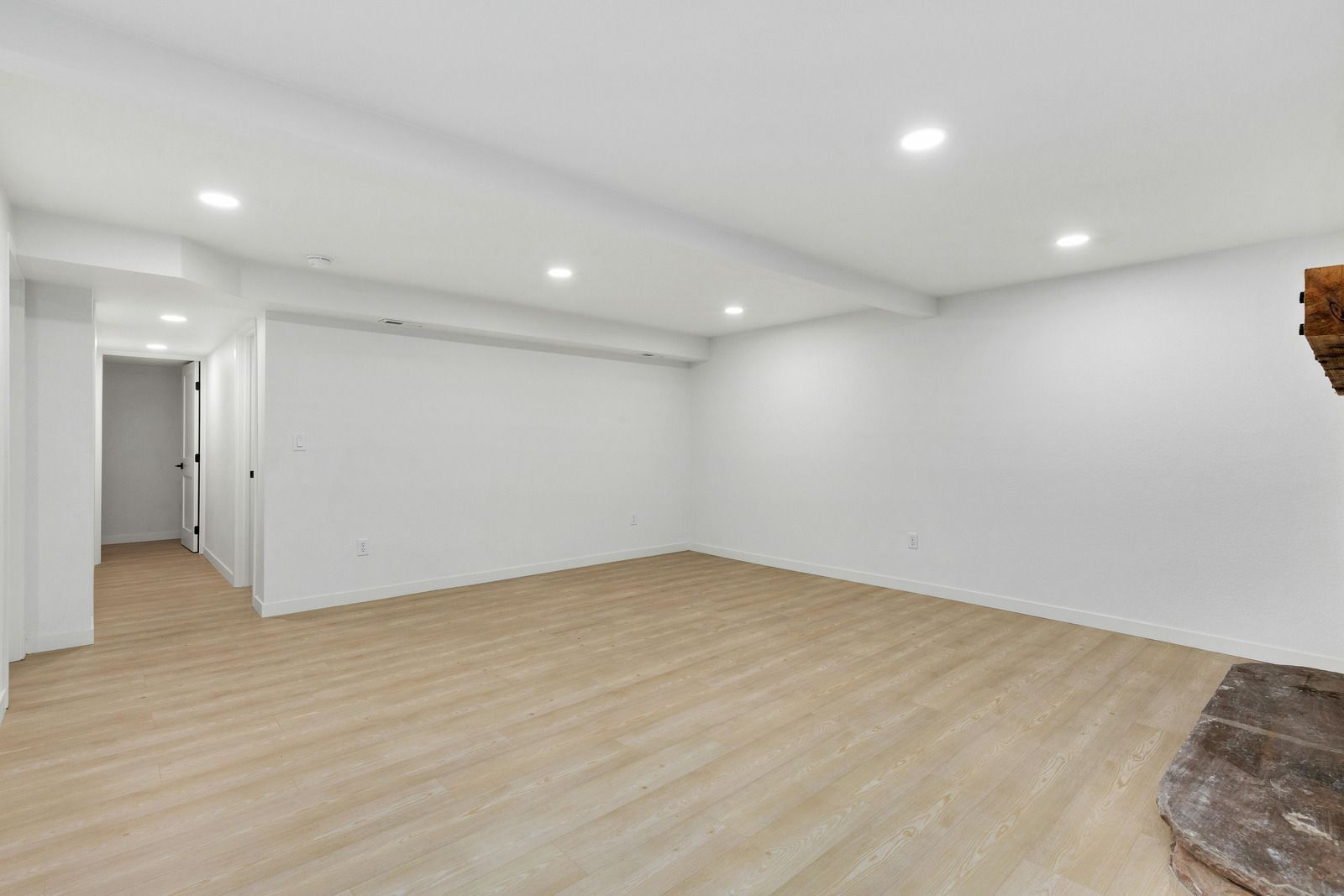 A modern, unfurnished room with white walls, recessed ceiling lights, light wood flooring, and a stone hearth feature.