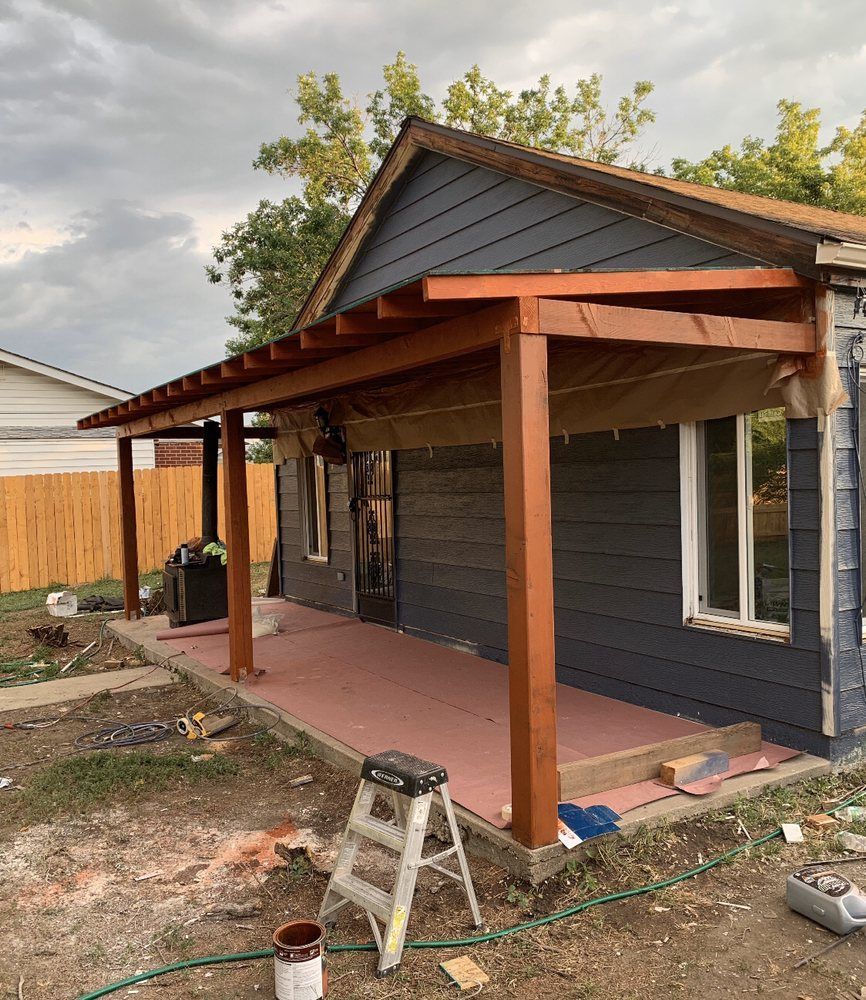 A wooden porch frame is under construction attached to a dark blue house with a concrete patio and a step ladder nearby.
