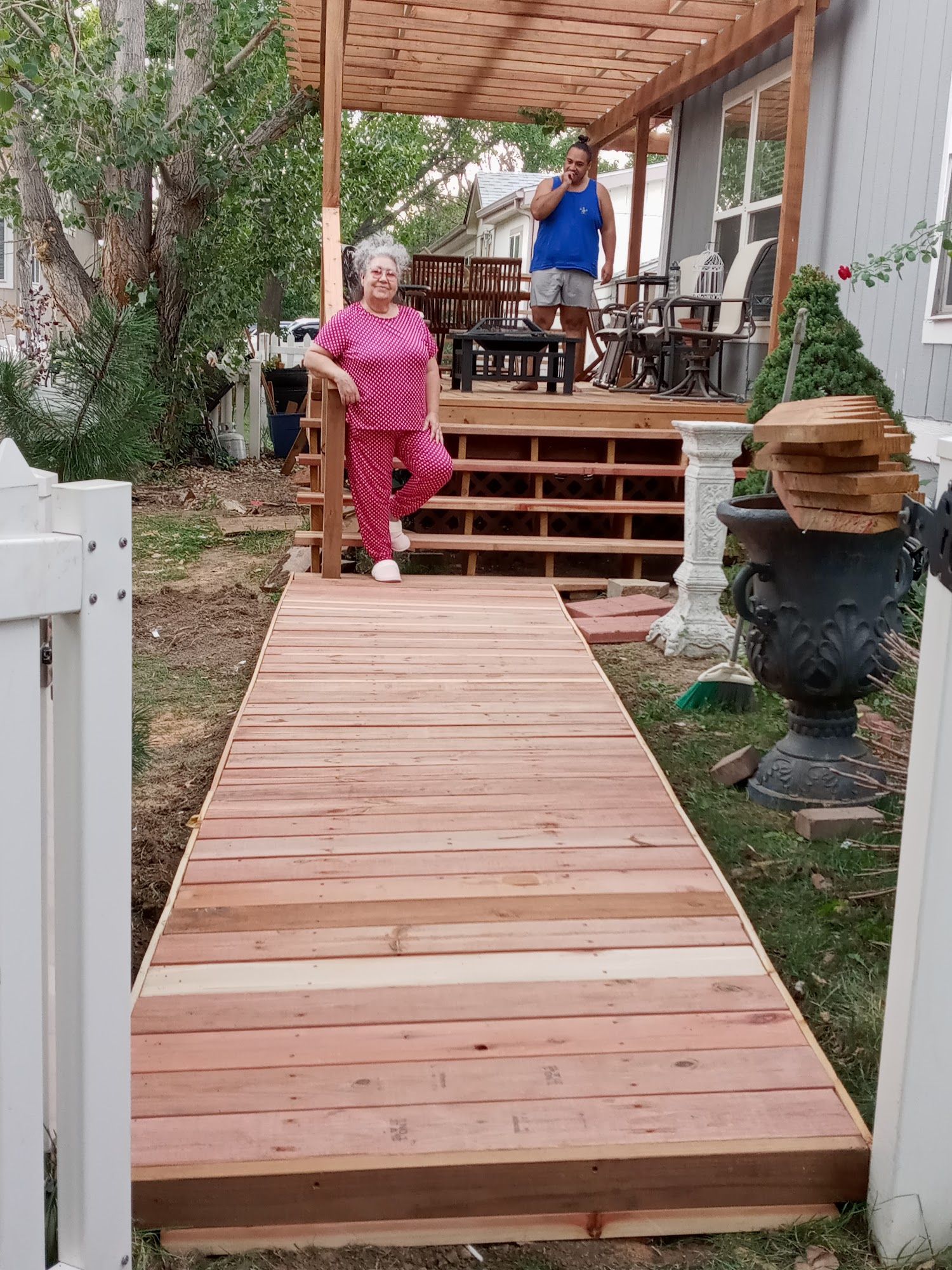 A person in a pink outfit stands on a new wooden walkway leading to a backyard deck with a pergola and another person.