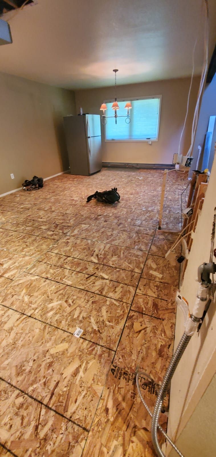 A room with bare plywood subflooring, a refrigerator in the corner, and an overhead light fixture near a window.