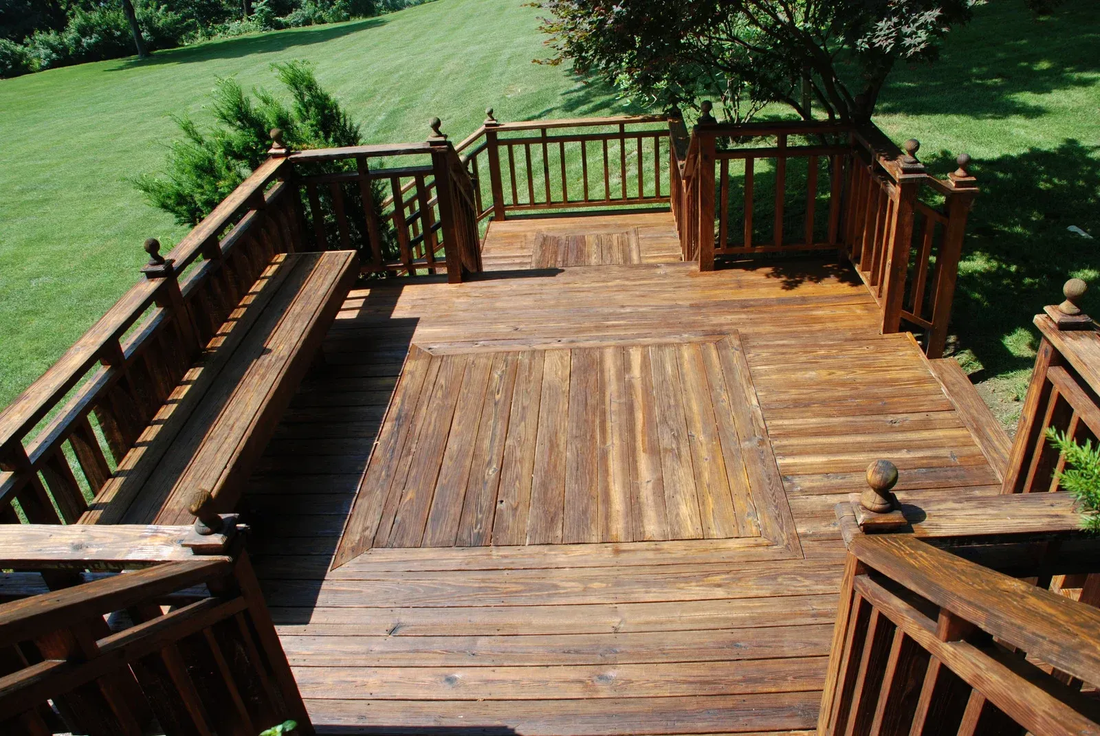 A stained wooden deck with built-in bench seating, railings, and steps, overlooking a grassy yard on a sunny day.