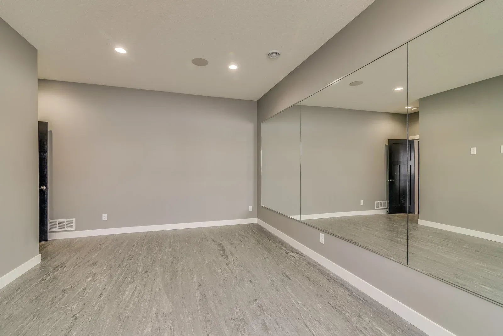 An empty fitness room with light grey walls, grey wood-look flooring, recessed lighting, and a large wall-to-wall mirror.