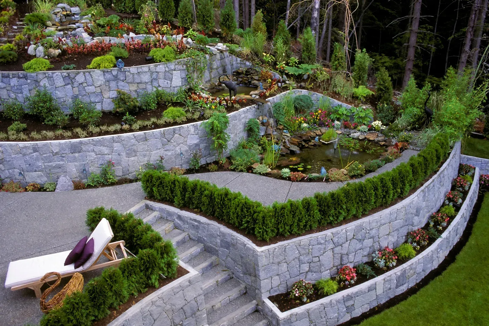 Tiered stone retaining walls create a garden landscape with a small pond, stairs, greenery, and a lounge chair.