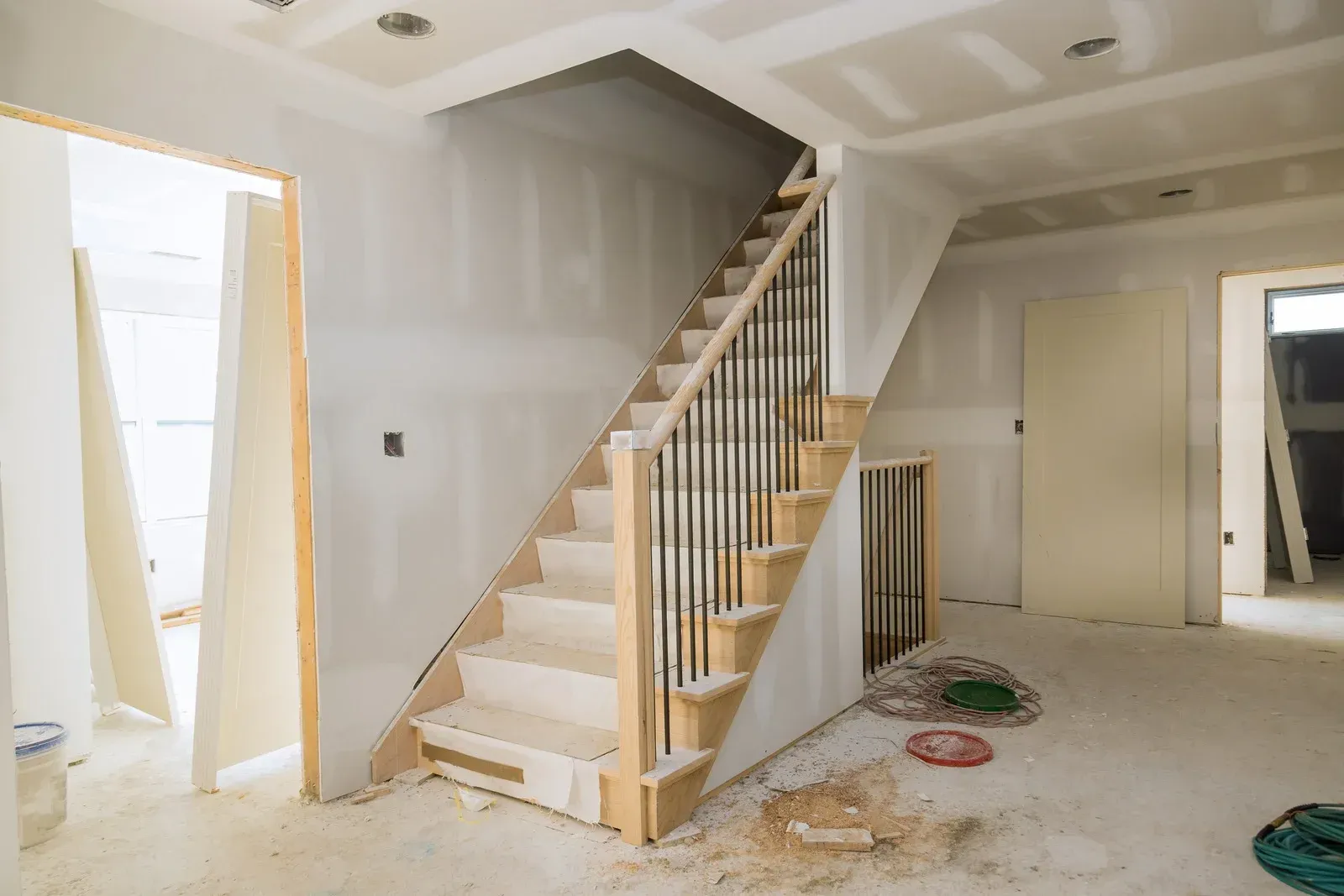 An interior room under construction features a wooden staircase with thin, dark metal balusters and unpainted drywall.
