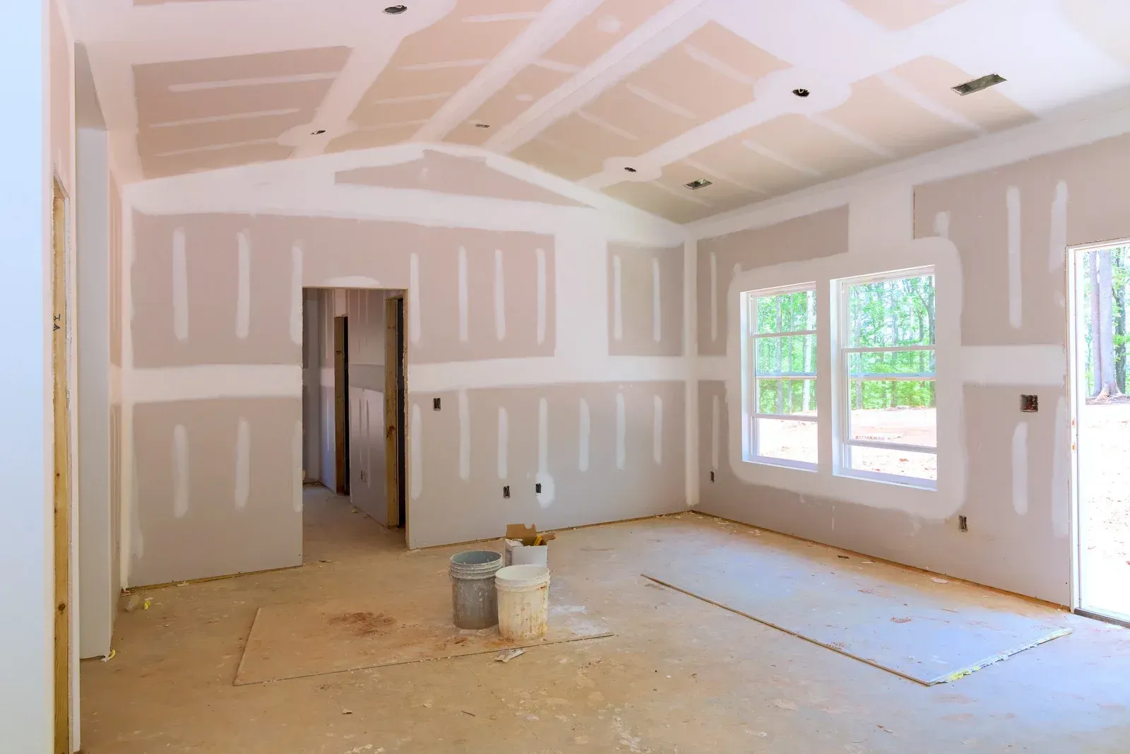 Interior of a room under construction with new drywall, taped seams, two large windows, and buckets on the floor.