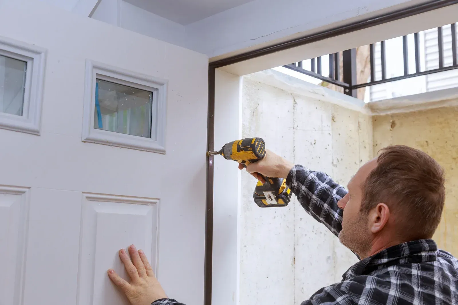A person in a plaid shirt uses a cordless drill to secure a white door into its frame.
