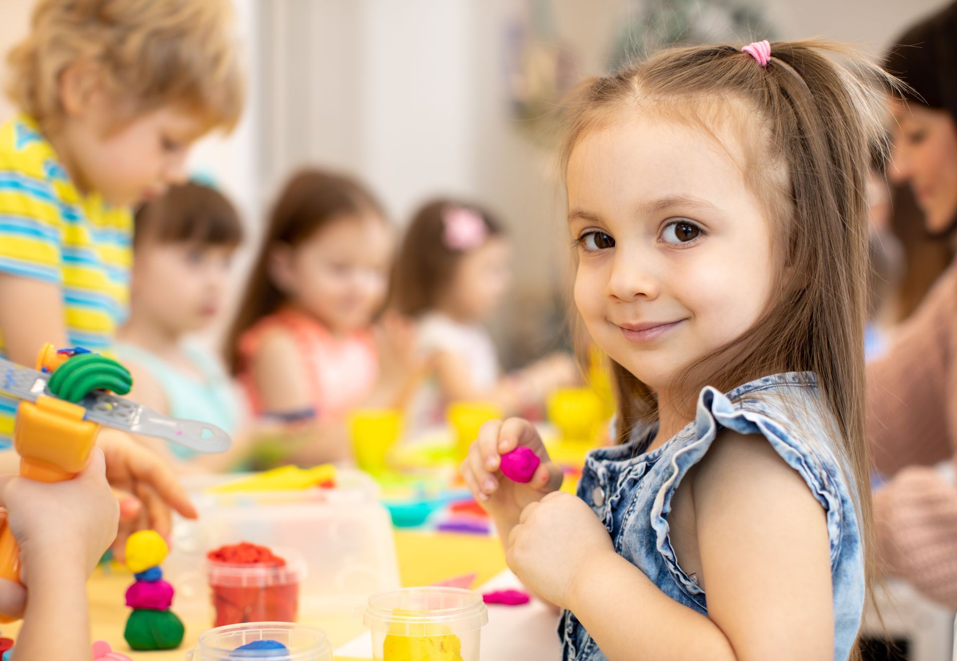 A happy little girl doing arts and crafts in a daycare centre with other children in the background.