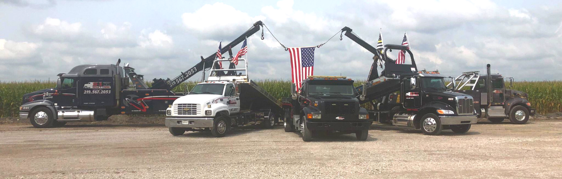Multiple tow trucks parked in a dirt lot with an American flag displayed. Green field and cloudy sky background.