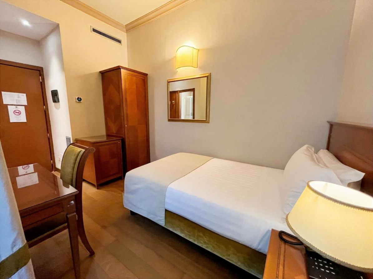 Habitación de hotel pequeña con dos camas individuales, armario, espejo y luz cálida gracias a una lámpara de pared.