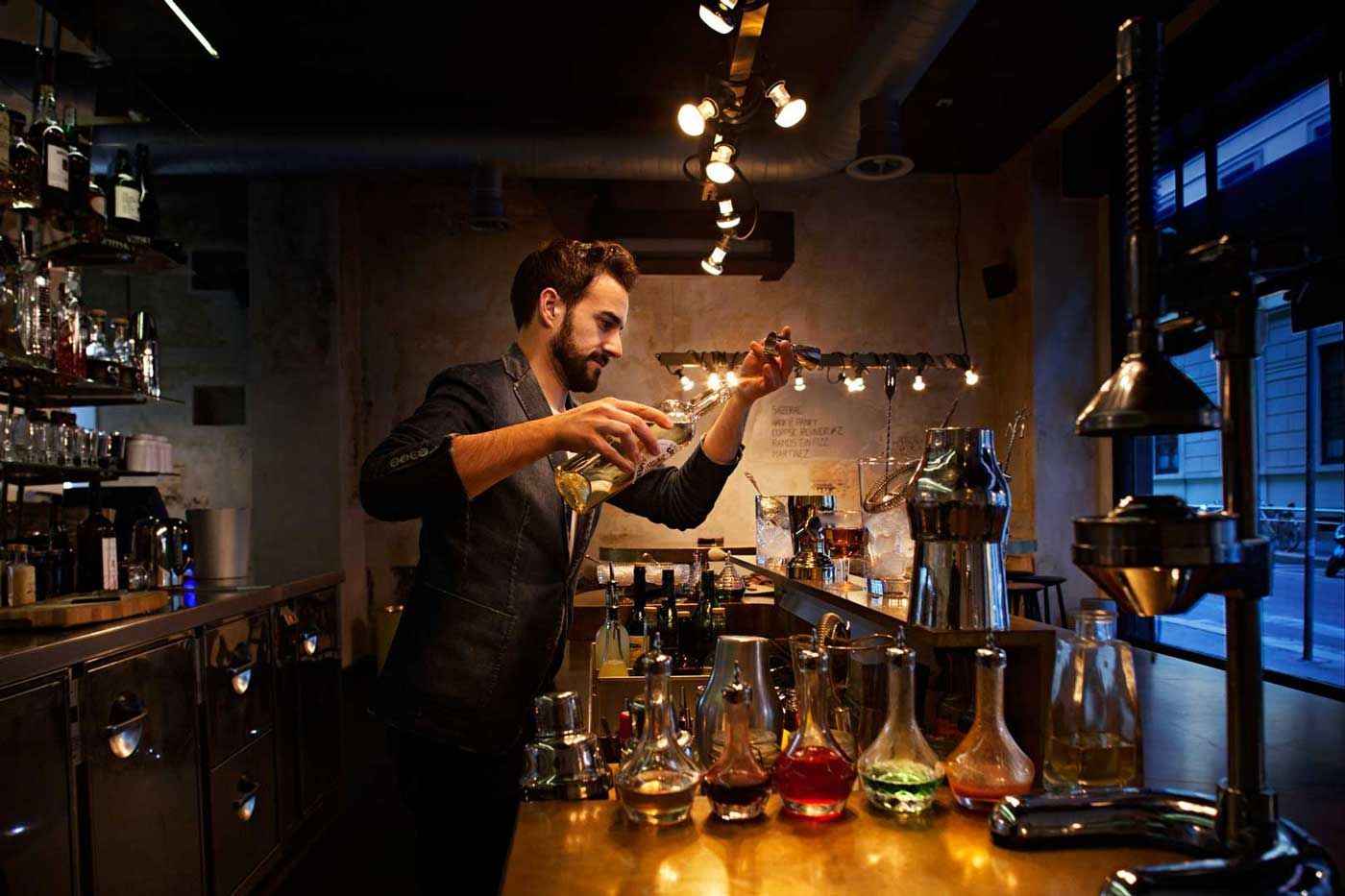 Un barista prepara un cocktail dietro un bancone scarsamente illuminato, utilizzando bottiglie e bicchieri.
