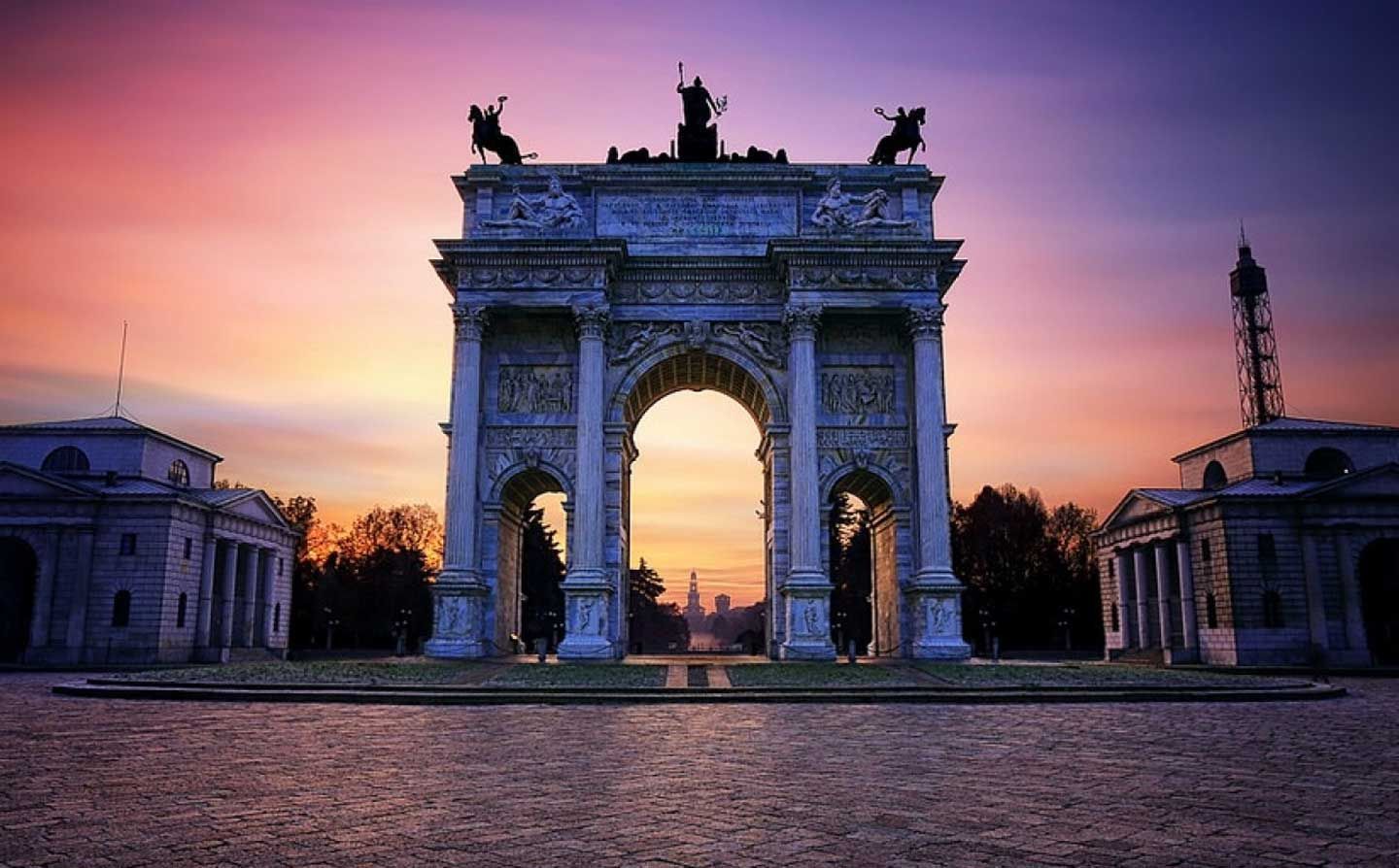 Un maestoso arco trionfale al tramonto, con le statue stagliate contro un cielo rosa-arancio.
