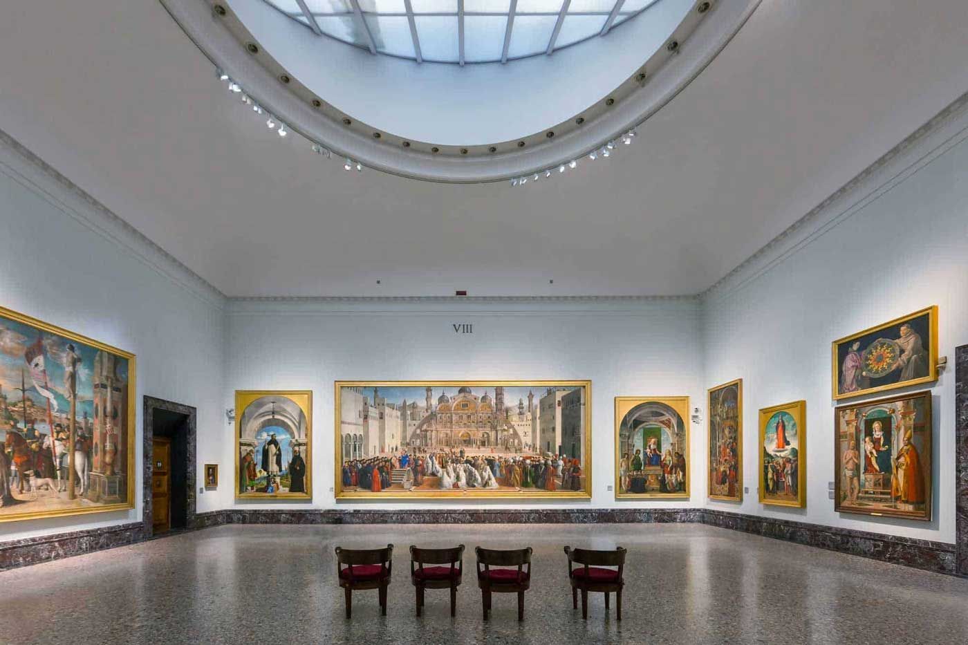 Luminosa galleria museale con quadri incorniciati su pareti bianche e tre sedie disposte al centro su un pavimento lucido.