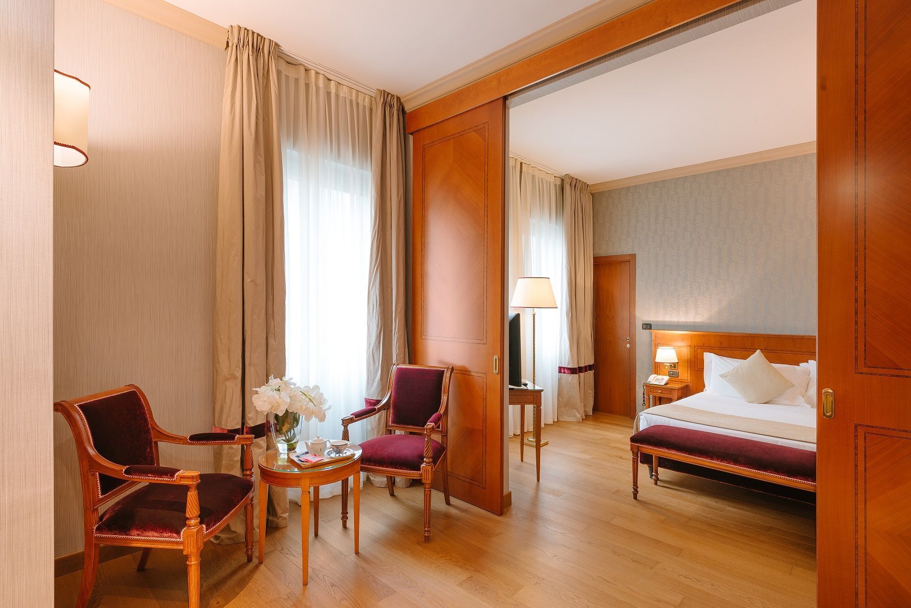 Habitación de hotel luminosa con cama, zona de estar con sillón y lámpara de pie, vista a través de puertas