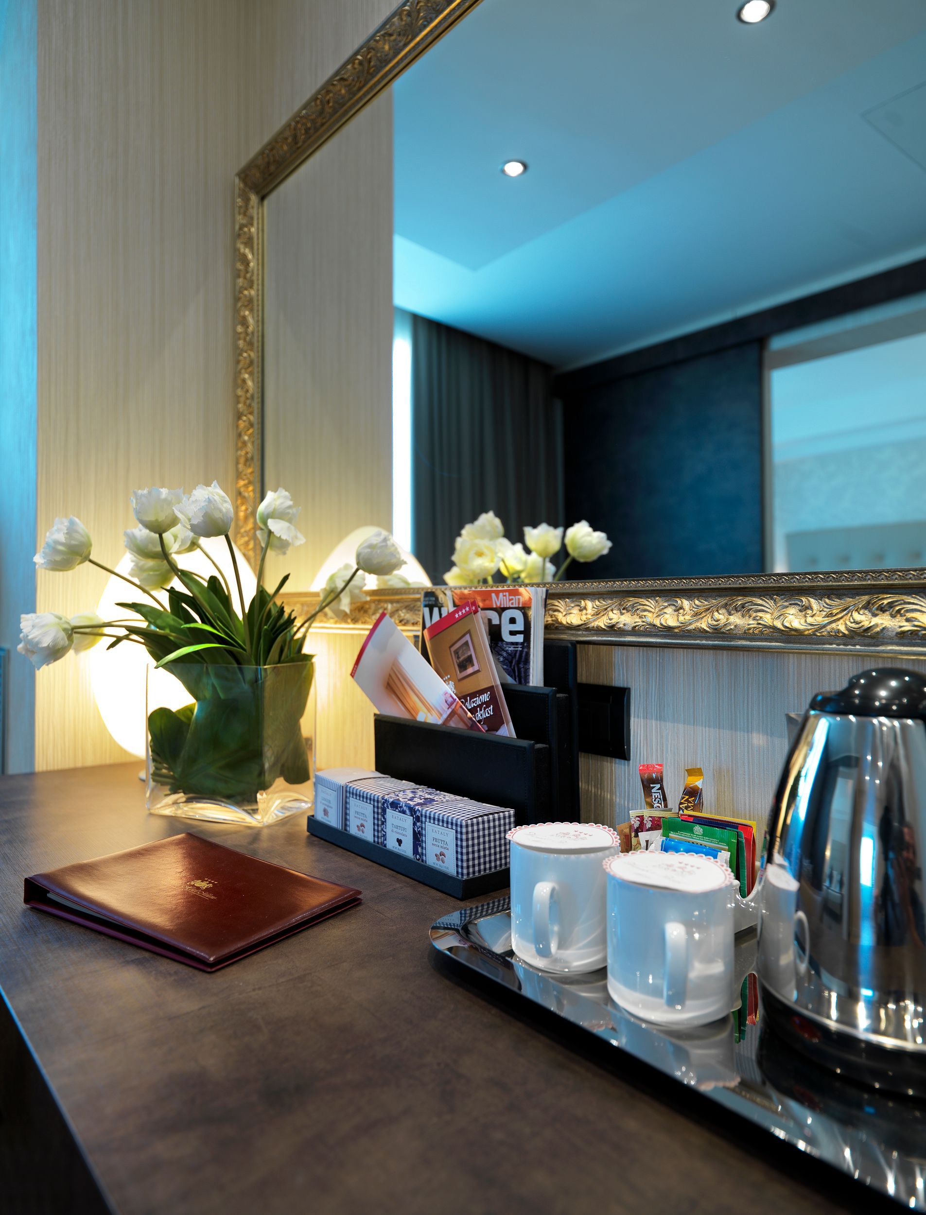 Mostrador de hotel con flores blancas, bandeja de café, tazas, hervidor de agua y un espejo con marco dorado.
