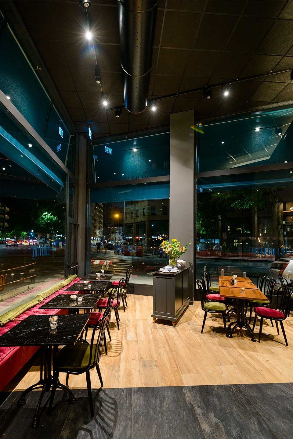 Interior de restaurante moderno con mesas, sillas, ventanales del suelo al techo y un ramo de flores en una mesa central.