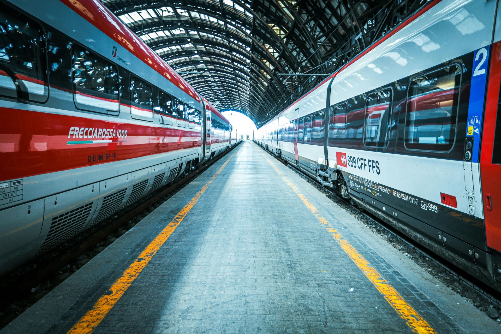 Andén vacío con dos trenes rojos y blancos bajo un moderno techo arqueado.