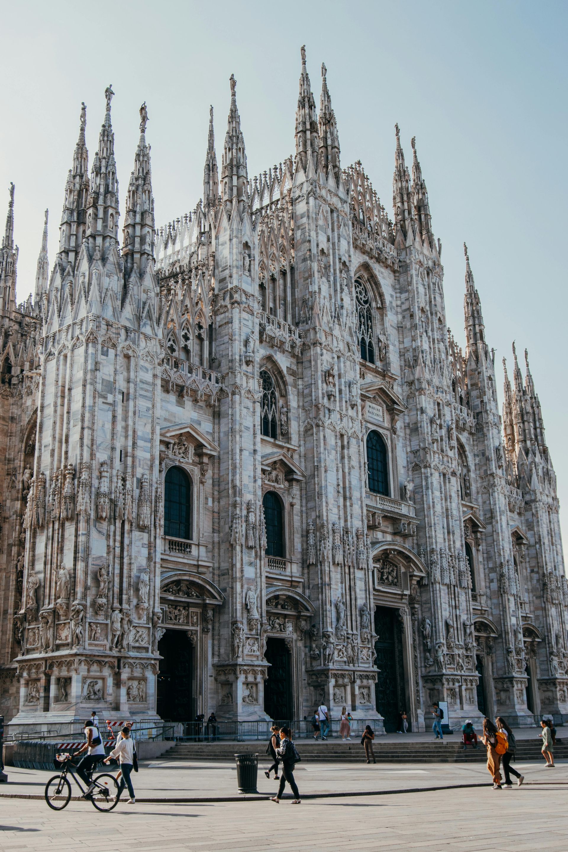 La ornamentada fachada gótica blanca de la catedral de Milán con algunas personas caminando por la plaza.