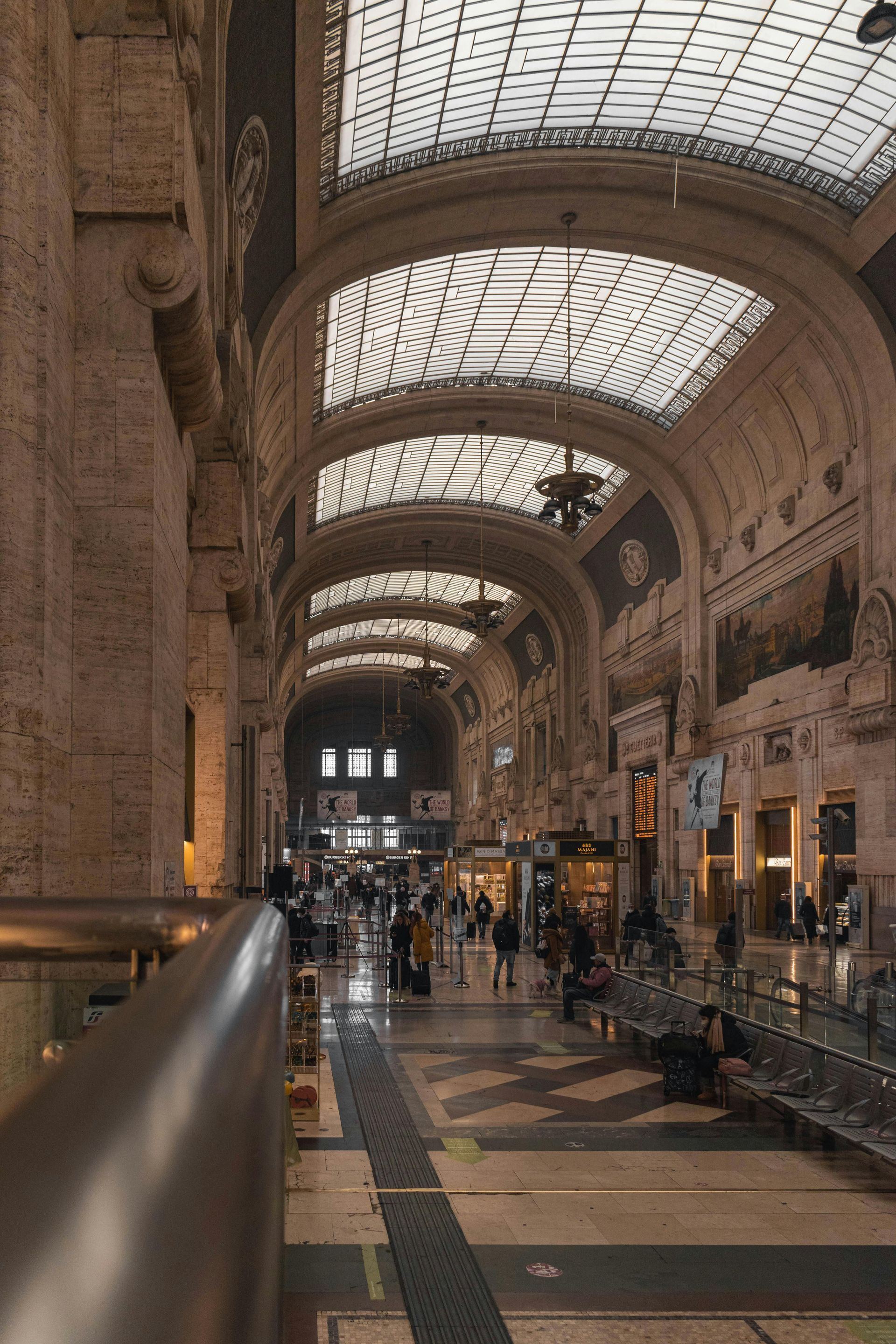 Gran vestíbulo de la estación de tren con arcos, techo de cristal, paredes de piedra ornamentadas y gente caminando abajo.