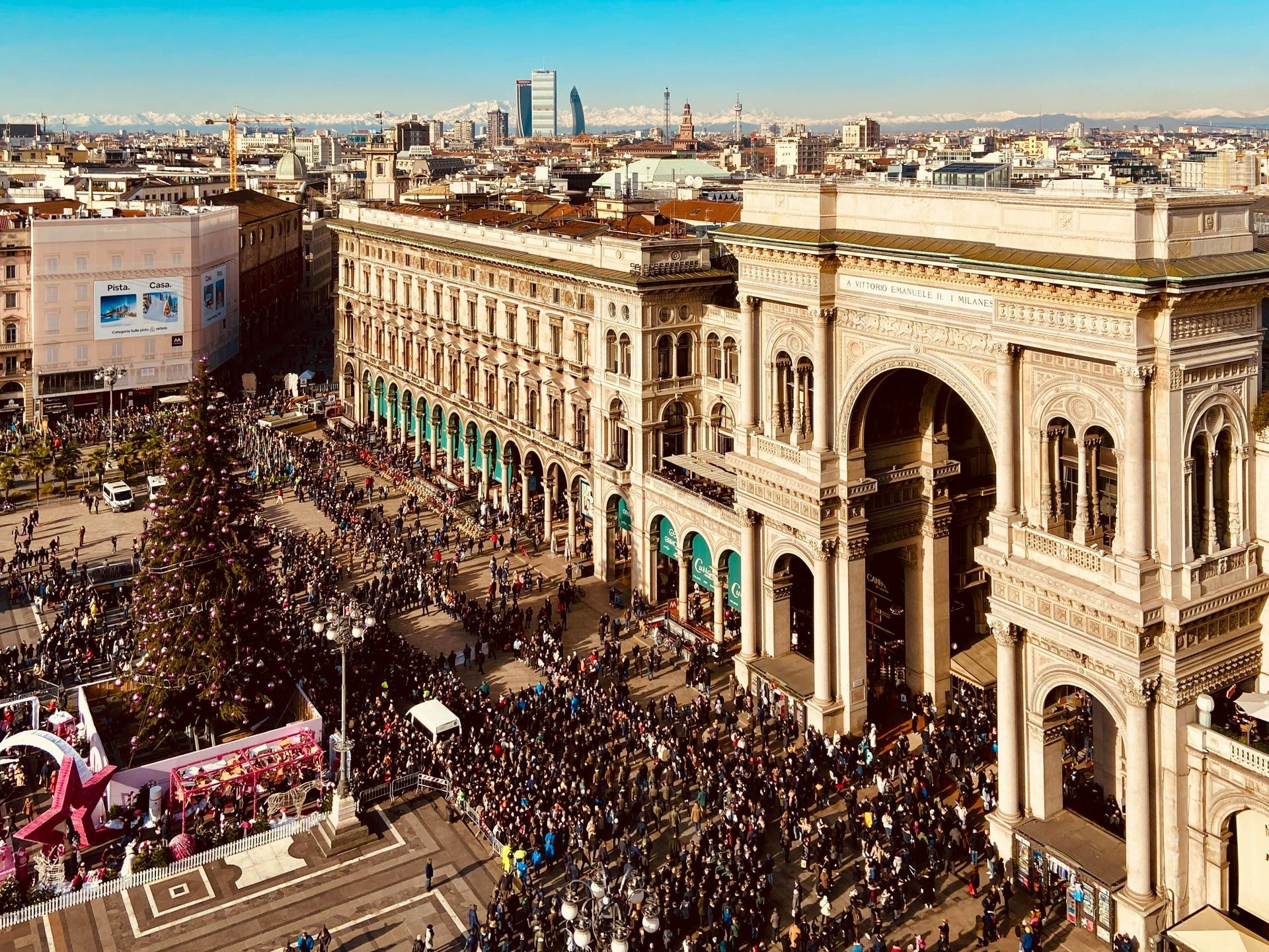Plaza de la ciudad abarrotada junto a edificios históricos ornamentados, con un desfile y un gran arco a plena luz del día.