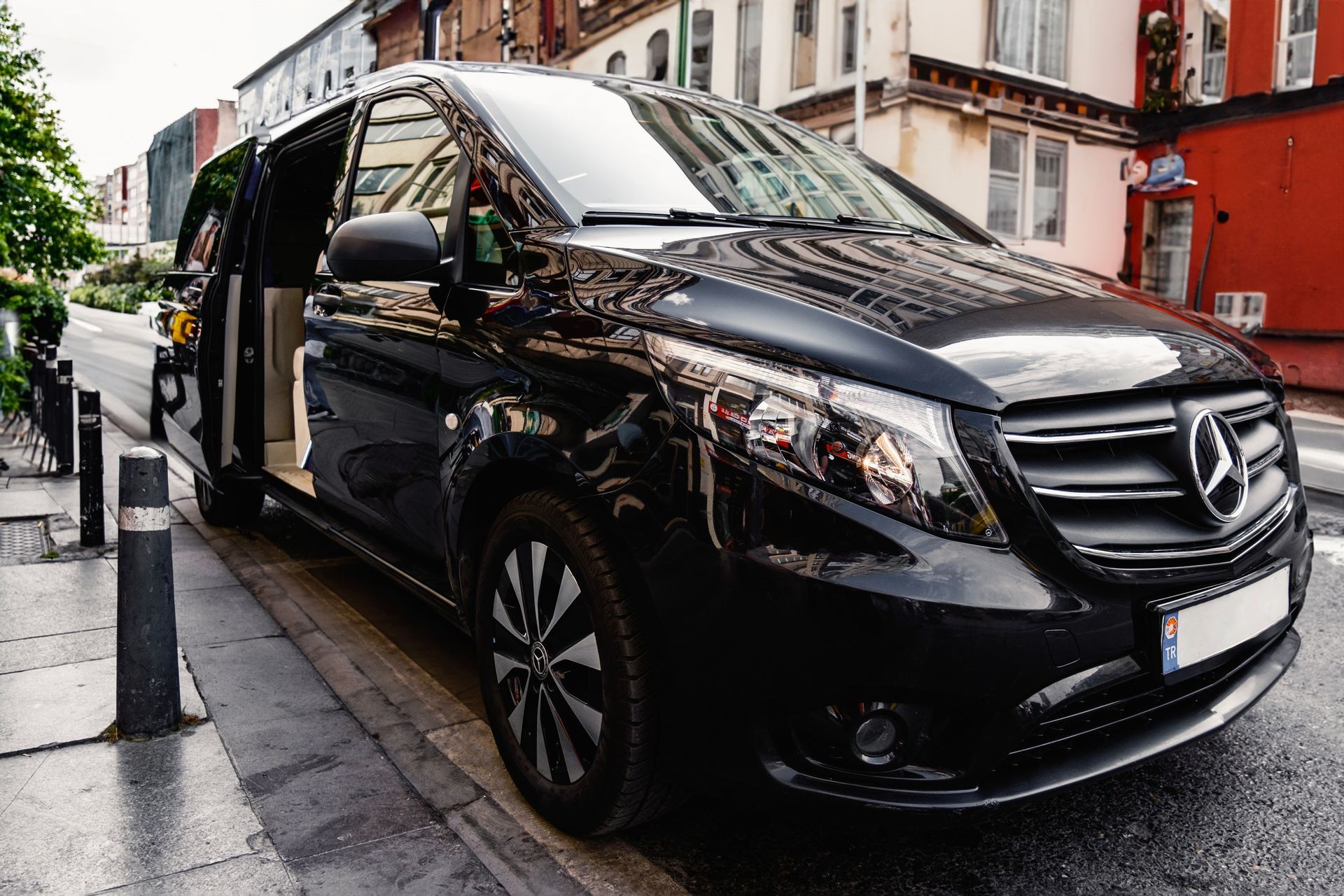 Una furgoneta Mercedes negra aparcada en una calle mojada de la ciudad, junto a edificios y escaparates rojos.