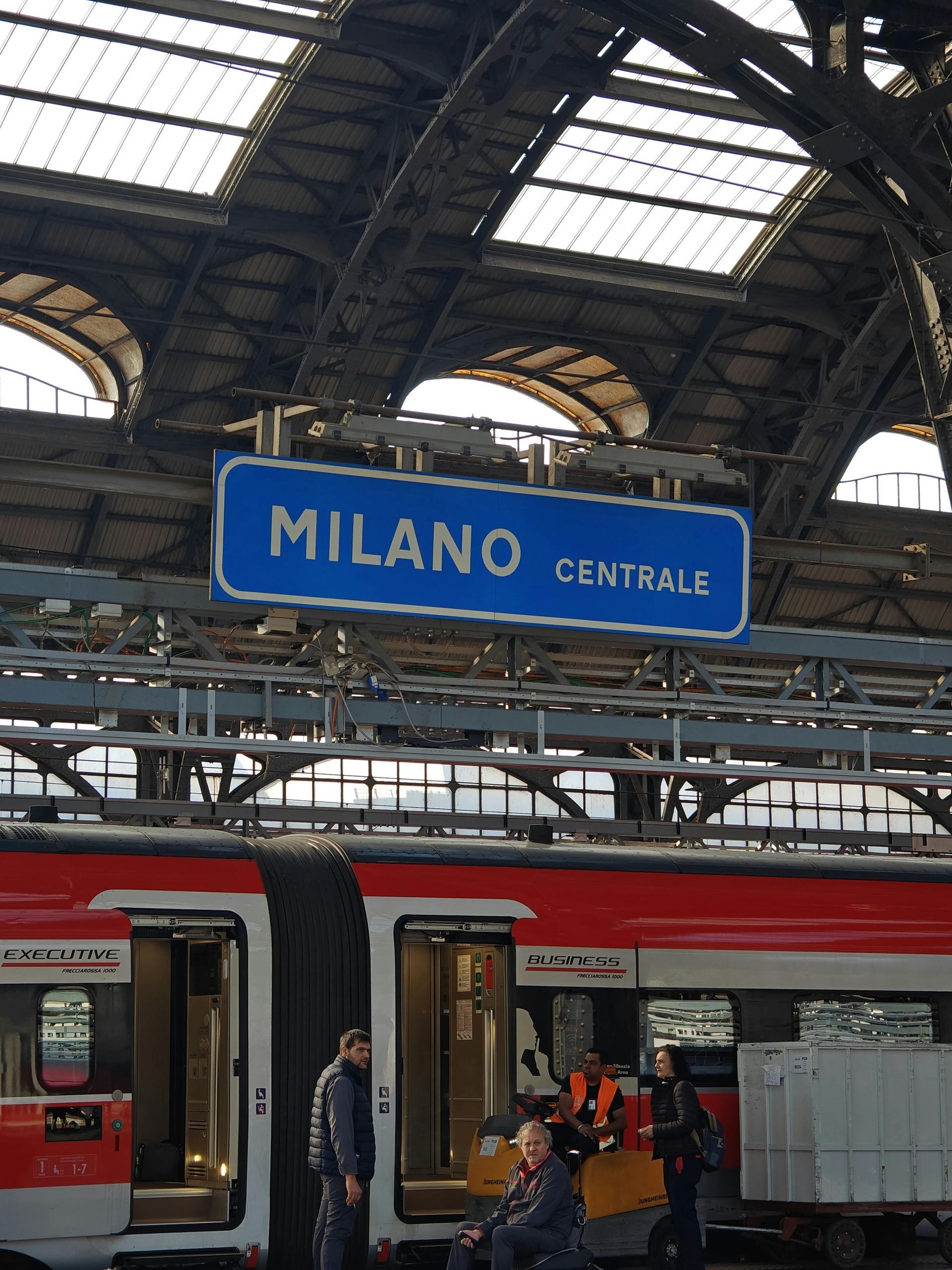 Letrero de la estación de tren Milano Centrale sobre un andén rojo con pasajeros y techo de cristal arqueado.
