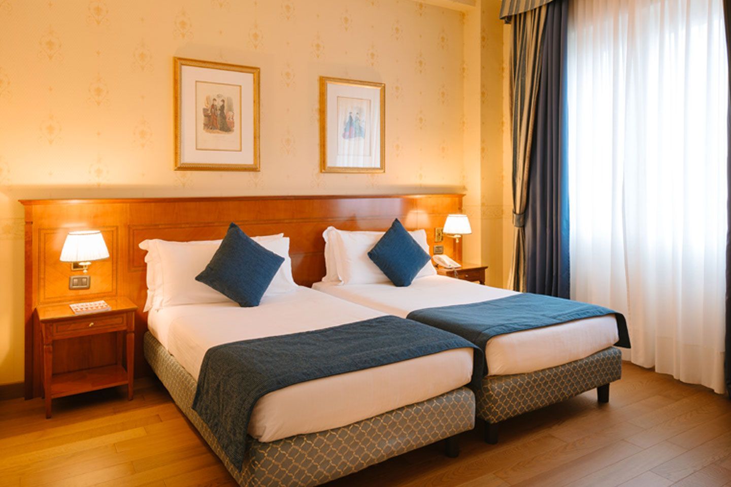 Acogedora habitación de hotel con dos camas individuales, detalles en azul, paneles de madera cálida y una ventana soleada.