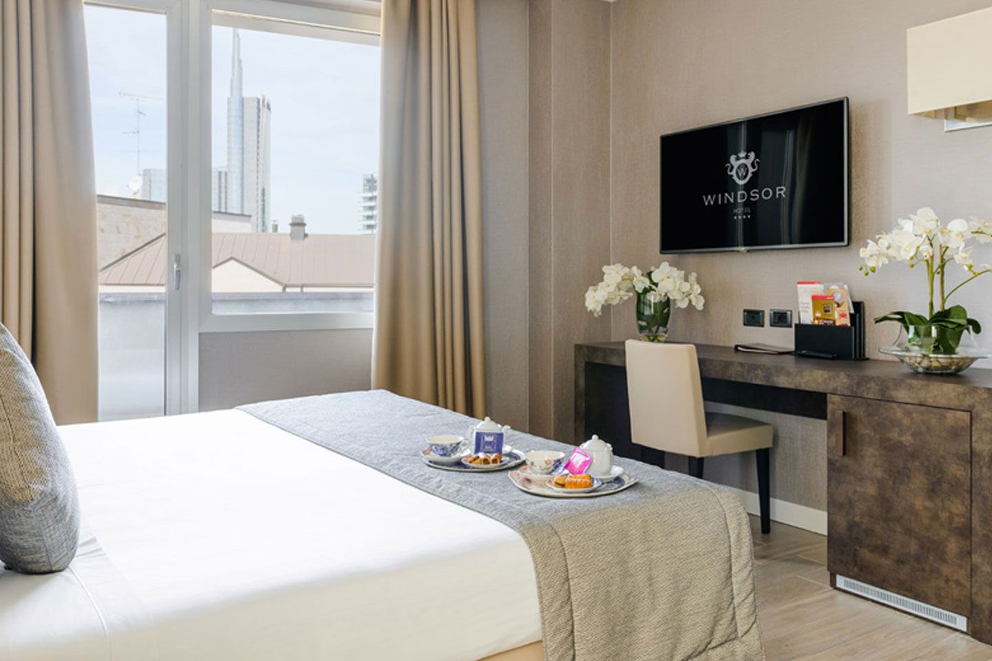 Habitación de hotel luminosa con cama, ventana, escritorio y TV, con flores y una bandeja de desayuno.