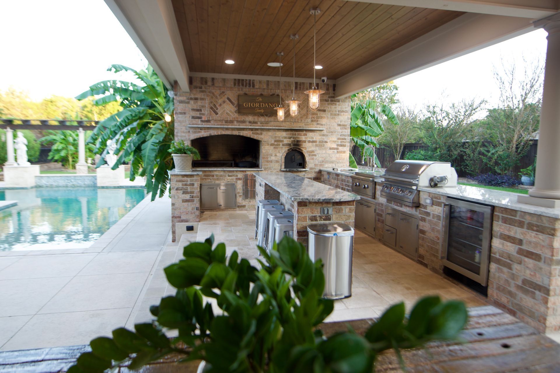 A large outdoor kitchen with a fireplace and a swimming pool in the background built by Fine Patio Design San Antonio TX